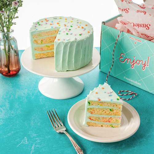 Image 2: Celebration 4-layer cake on a table with a slice of the cake next to it. The cake and slice show the confetti baked into the vanilla cake and the SusieBlue (light blue) buttercream. In the background you see the shipping box that the cake is shipped it. View 2