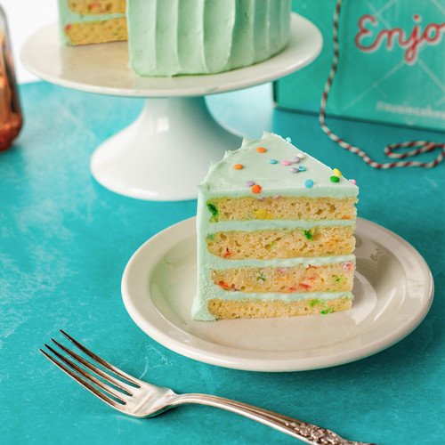Image 3: Celebration 4-layer cake slice on the table with a fork ready to be enjoyed. The slice gives a close up visual of the cake with colorful confetti baked into the vanilla cake and the SusieBlue (light blue) buttercream. view 3