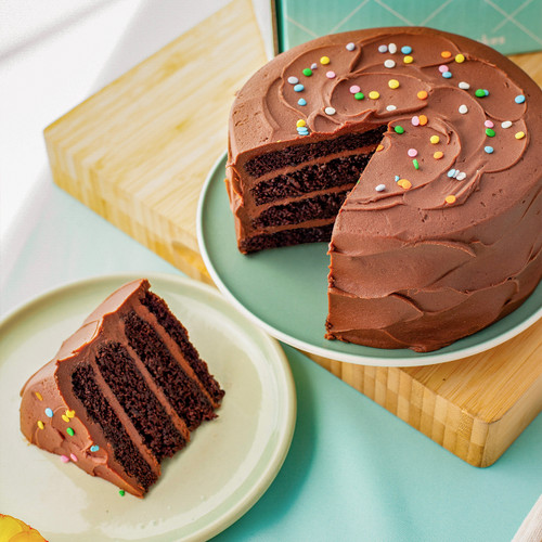 Image 3: Chocolate cake shown from a top down view showing the chocolate cake topped with colorful confetti sprinkles. The slice of cake is shown next to the table so you can visually see the 4 layers of moist chocolate cake with the chocolate buttercream. view 3