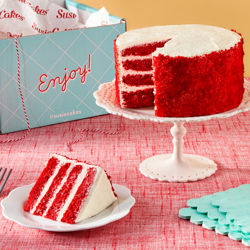 Red velvet cake with a slice next to it on a table. In the background you see the shipping box the cake arrived in. View 2